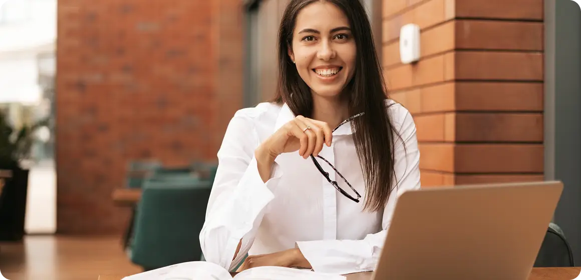 Professora sorrindo segurando os óculos em frente ao notebook